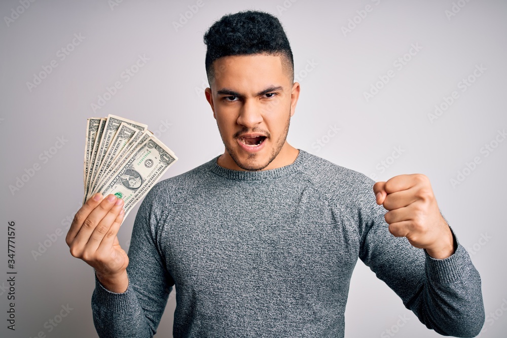 Young handsome man holding bunch of dollars banknotes over isolated white background annoyed and frustrated shouting with anger, crazy and yelling with raised hand, anger concept Young handsome man holding bunch of dollars banknotes over isolated white background annoyed and frustrated shouting with anger, crazy and yelling with raised hand, anger concept
