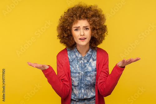 How could you? Portrait of annoyed woman with fluffy curly hair in casual outfit raising hands in indignant gesture, quarrelling, having conflict. indoor studio shot isolated on yellow background