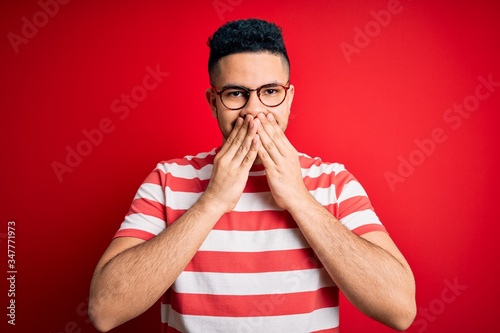 Young handsome man wearing casual striped t-shirt and glasses over isolated red background laughing and embarrassed giggle covering mouth with hands, gossip and scandal concept