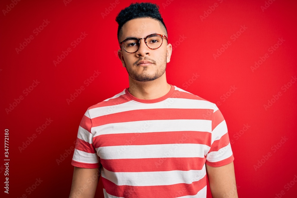 Young handsome man wearing casual striped t-shirt and glasses over isolated red background Relaxed with serious expression on face. Simple and natural looking at the camera. Young handsome man wearing casual striped t-shirt and glasses over isolated red background Relaxed with serious expression on face. Simple and natural looking at the camera.