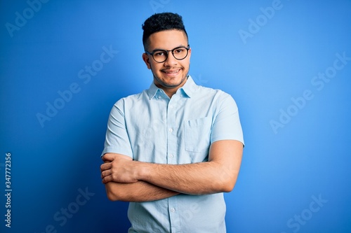 Young handsome man wearing casual summer shirt and glasses over isolated blue background happy face smiling with crossed arms looking at the camera. Positive person.