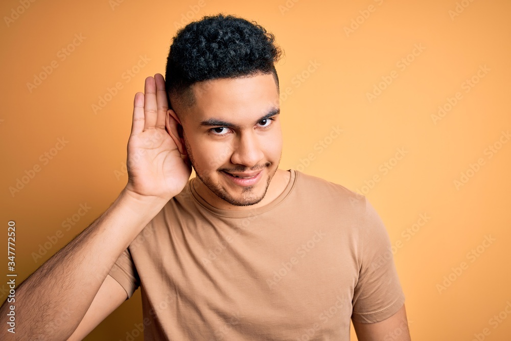 Young handsome man wearing casual t-shirt standing over isolated yellow background smiling with hand over ear listening an hearing to rumor or gossip. Deafness concept. Young handsome man wearing casual t-shirt standing over isolated yellow background smiling with hand over ear listening an hearing to rumor or gossip. Deafness concept.