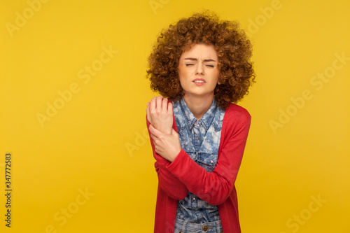 Injured hand. Portrait of young curly-haired woman wincing in pain and touching sore wrist  suffering joint inflammation of rheumatoid arthritis  stiffness. studio shot isolated on yellow background