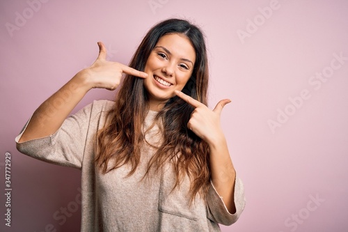 Young beautiful brunette woman wearing casual sweater standing over pink background smiling cheerful showing and pointing with fingers teeth and mouth. Dental health concept.