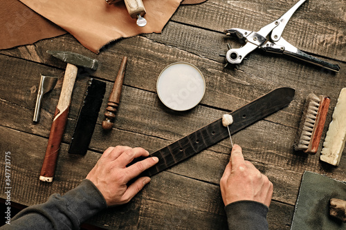 Working process of the leather belt in leather workshop. Man holding tool. Tanner in old tannery. Wooden table background. Close up man arm. Maintenance concept. Goods production. Top view, flat lay.