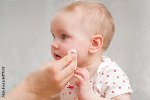 Young mother hand wiping baby girl cheek with white cotton pad. Daily routine. Care about infant clean and soft body skin. Closeup. Side view. photo