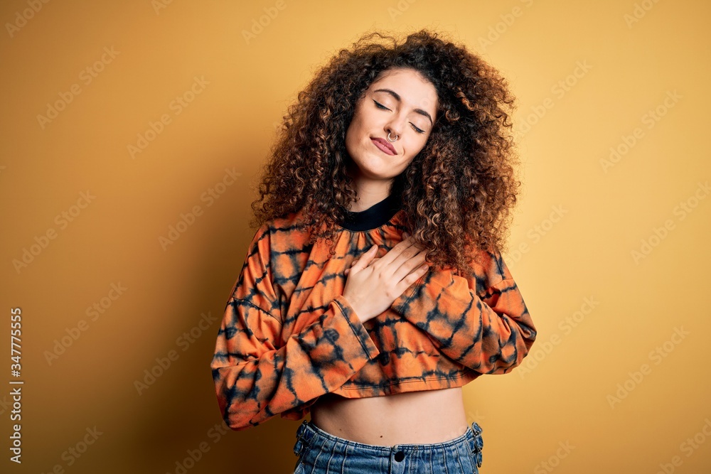 Young beautiful brunette woman with curly hair and piercing wearing casual t-shirt smiling with hands on chest with closed eyes and grateful gesture on face. Health concept. Young beautiful brunette woman with curly hair and piercing wearing casual t-shirt smiling with hands on chest with closed eyes and grateful gesture on face. Health concept.