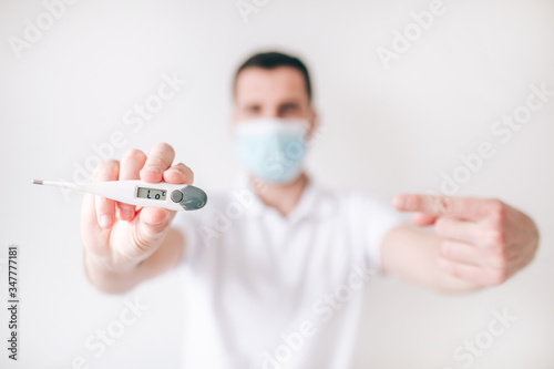 Young sick man isolated over white background. Guy hold thermometer in hand and point on it with finger. High temperature. Sick ill person wear facial medical mask.