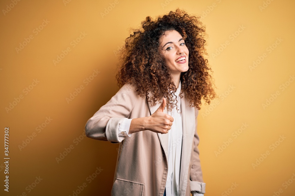 Young beautiful brunette woman with curly hair and piercing wearing casual t-shirt and diadem doing happy thumbs up gesture with hand. Approving expression looking at the camera showing success. Young beautiful brunette woman with curly hair and piercing wearing casual t-shirt and diadem doing happy thumbs up gesture with hand. Approving expression looking at the camera showing success.
