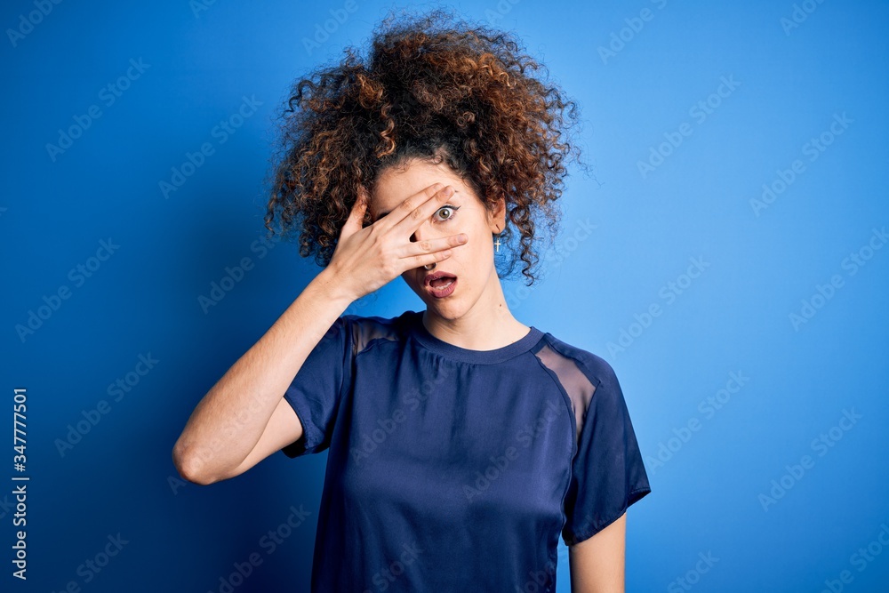 Young beautiful woman with curly hair and piercing wearing casual blue t-shirt peeking in shock covering face and eyes with hand, looking through fingers with embarrassed expression. Young beautiful woman with curly hair and piercing wearing casual blue t-shirt peeking in shock covering face and eyes with hand, looking through fingers with embarrassed expression.