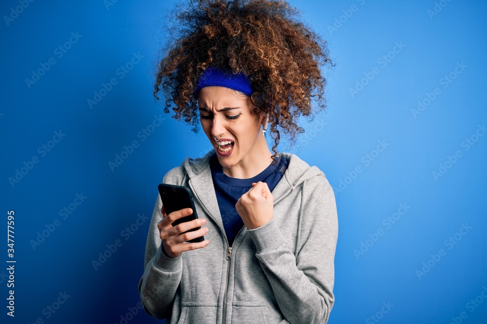 Beautiful sportswoman with curly hair and piercing wearing sportswear using smartphone annoyed and frustrated shouting with anger, crazy and yelling with raised hand, anger concept Beautiful sportswoman with curly hair and piercing wearing sportswear using smartphone annoyed and frustrated shouting with anger, crazy and yelling with raised hand, anger concept