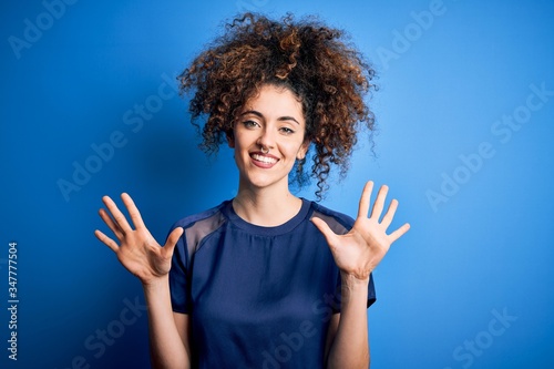 Young beautiful woman with curly hair and piercing wearing casual blue t-shirt showing and pointing up with fingers number ten while smiling confident and happy.