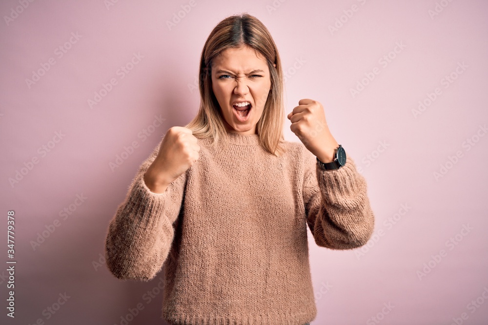 Young beautiful blonde woman wearing winter wool sweater over pink isolated background angry and mad raising fists frustrated and furious while shouting with anger. Rage and aggressive concept. Young beautiful blonde woman wearing winter wool sweater over pink isolated background angry and mad raising fists frustrated and furious while shouting with anger. Rage and aggressive concept.
