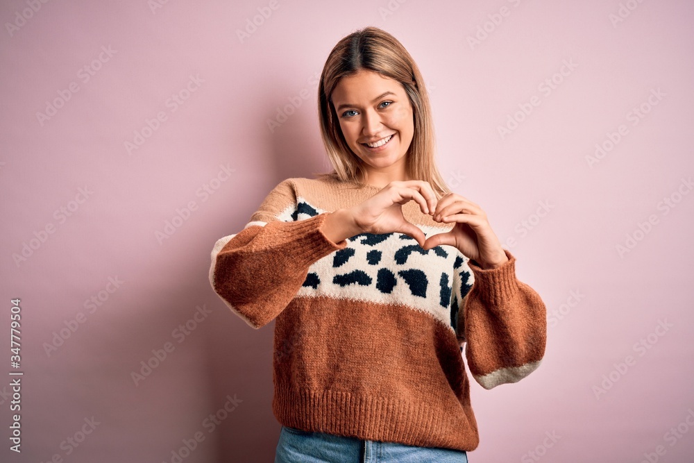 Young beautiful blonde woman wearing fashion animal print sweater over pink background smiling in love showing heart symbol and shape with hands. Romantic concept. Young beautiful blonde woman wearing fashion animal print sweater over pink background smiling in love showing heart symbol and shape with hands. Romantic concept.