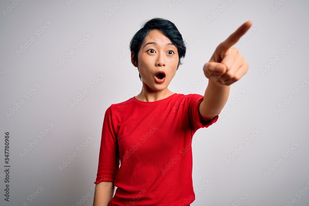 Young beautiful asian girl wearing casual red t-shirt standing over isolated white background Pointing with finger surprised ahead, open mouth amazed expression, something on the front Young beautiful asian girl wearing casual red t-shirt standing over isolated white background Pointing with finger surprised ahead, open mouth amazed expression, something on the front