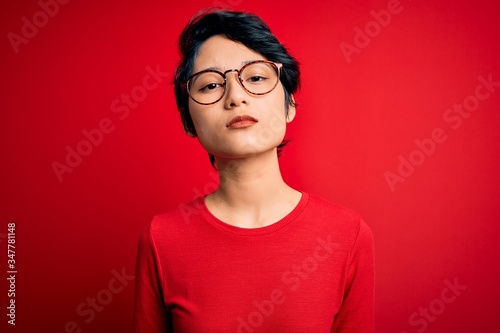 Young beautiful asian girl wearing casual t-shirt and glasses over isolated red background Relaxed with serious expression on face. Simple and natural looking at the camera. © Krakenimages.com