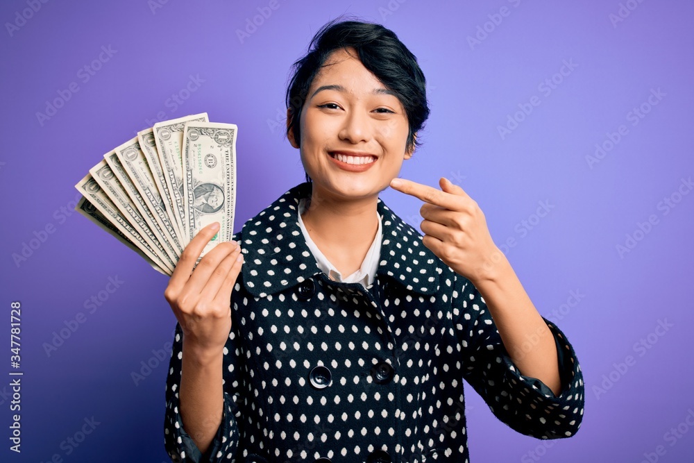 Young beautiful asian girl holding bunch of dollars banknotes over purple background very happy pointing with hand and finger Young beautiful asian girl holding bunch of dollars banknotes over purple background very happy pointing with hand and finger