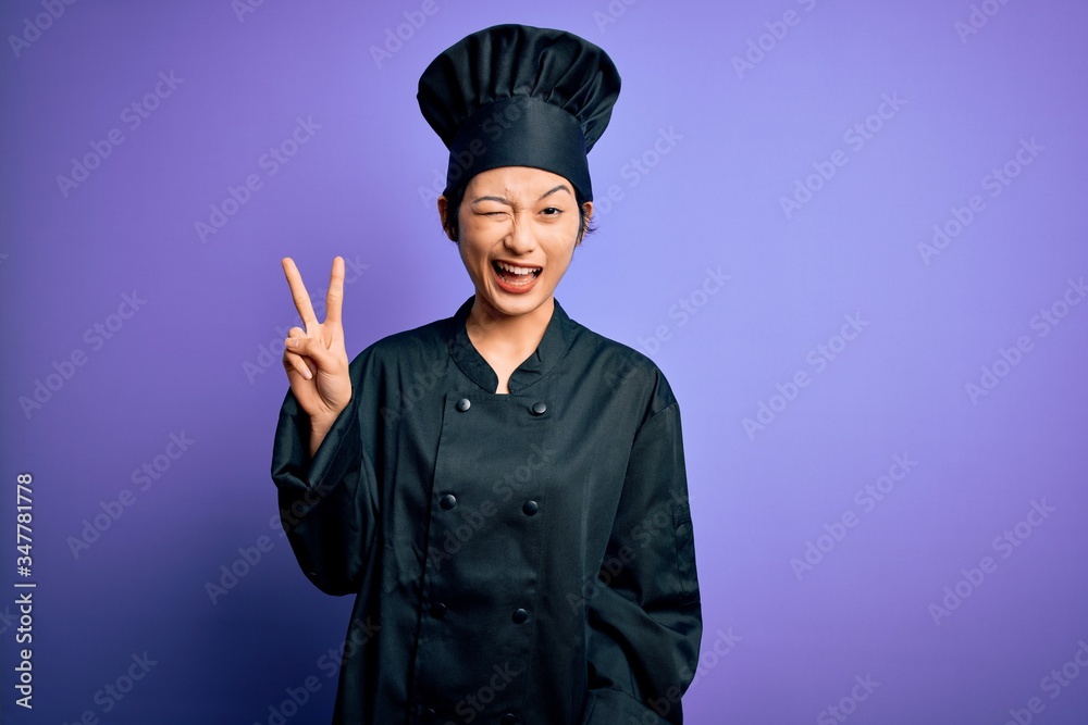 Young beautiful chinese chef woman wearing cooker uniform and hat over purple background smiling with happy face winking at the camera doing victory sign with fingers. Number two. Young beautiful chinese chef woman wearing cooker uniform and hat over purple background smiling with happy face winking at the camera doing victory sign with fingers. Number two.