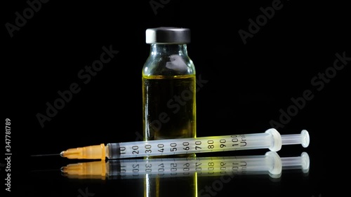 Glass vial filled with yellow liquid vaccine and insulin syringe rotate on a black background. Pharmaceutical industry. Viral infection treatment.