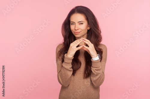 Portrait of scheming devious attractive young woman with brunette hair clasping hands and thinking over tricky plan, having sly cunning idea to prank. indoor studio shot isolated on pink background