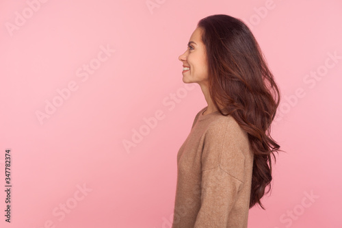 Side view of happy young woman with long brunette wavy hair smiling joyfully, expressing optimism, positive emotions, female beauty and fashion concept. indoor studio shot isolated on pink background © khosrork