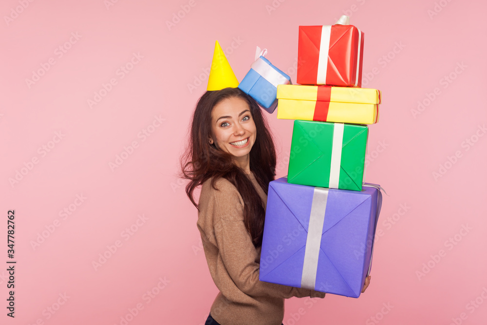 Fototapeta premium Portrait of excited joyful young woman wearing party cone and smiling pleased as holding mount of gift boxes, enjoying many best presents, celebrating birthday. studio shot isolated on pink background