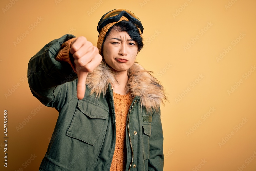 Beautiful asian skier girl wearing snow sportswear using ski goggles over yellow background looking unhappy and angry showing rejection and negative with thumbs down gesture. Bad expression. Beautiful asian skier girl wearing snow sportswear using ski goggles over yellow background looking unhappy and angry showing rejection and negative with thumbs down gesture. Bad expression.