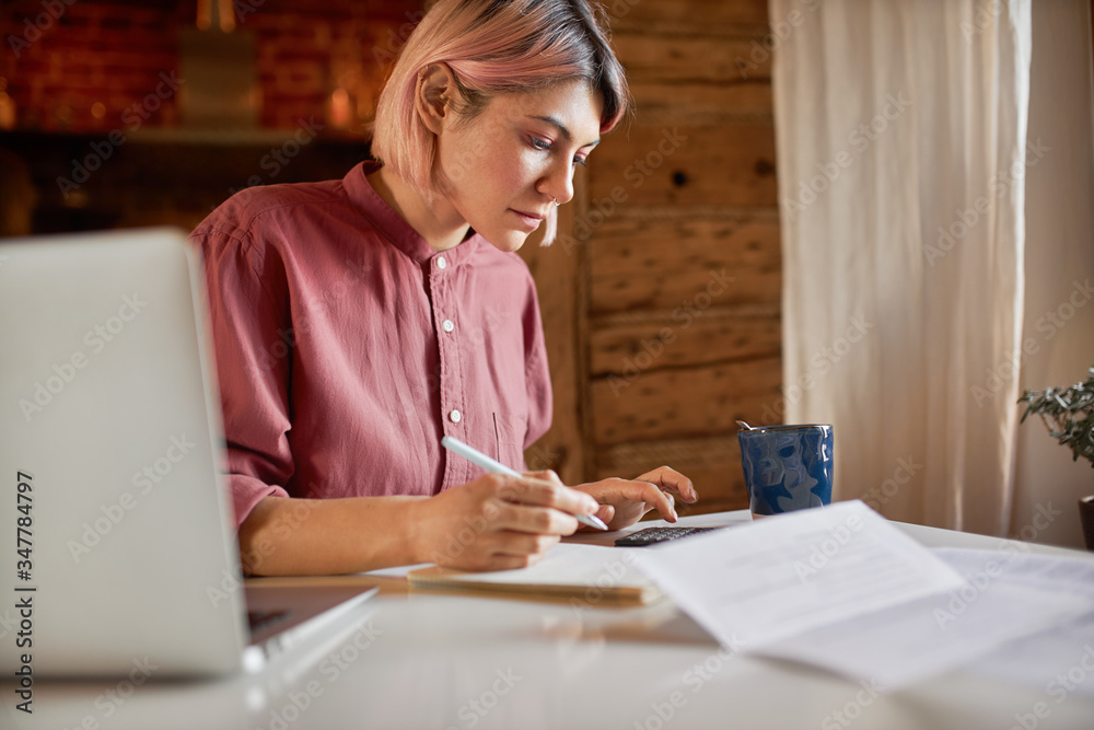 Side view of serious young female outsourced bookeeper providing accounting service for small businesses, sitting at desk with portable computer, holding pen to make notes and using calculator Side view of serious young female outsourced bookeeper providing accounting service for small businesses, sitting at desk with portable computer, holding pen to make notes and using calculator