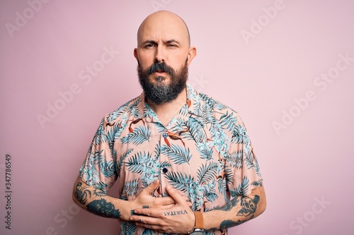 Handsome bald man with beard and tattoo wearing casual floral shirt over pink background with hand on stomach because indigestion, painful illness feeling unwell. Ache concept.