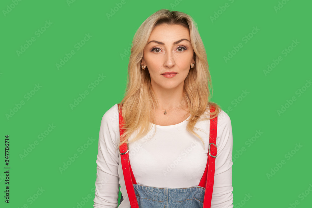 Portrait of serious concentrated adult woman with blond hair in stylish denim overalls looking at camera with focused calm pensive face expression. indoor studio shot isolated on green background