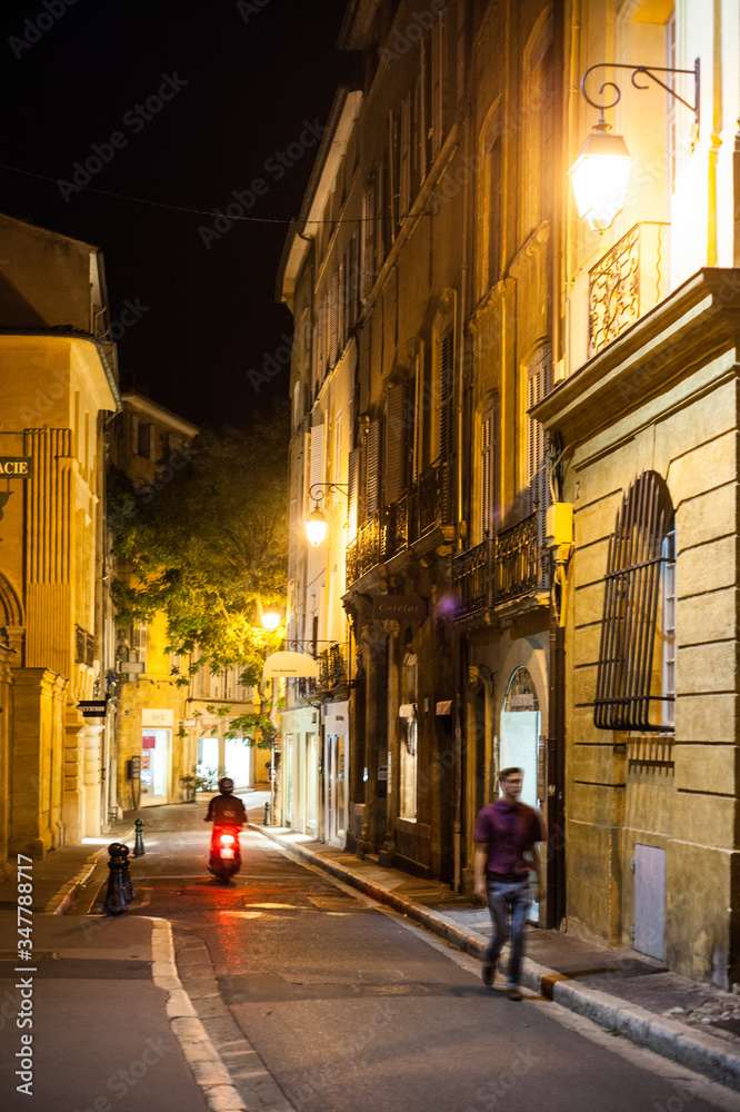 Aix-en-Provence, France - Vue de nuit d'une rue de la vieille ville avec un passant qui marche et un scooter qui roule.