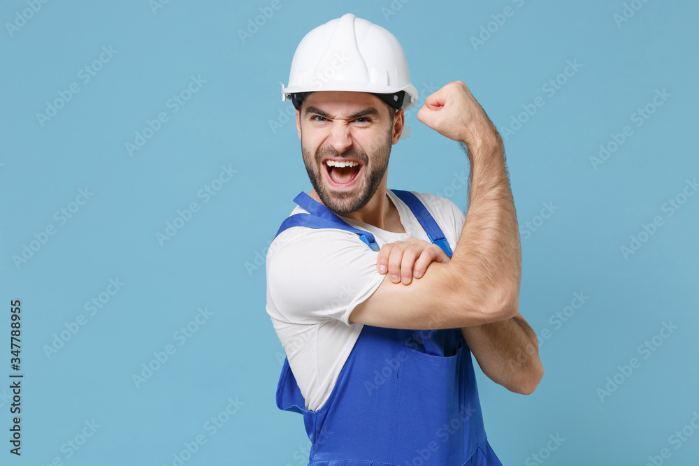 Funny young man in coveralls protective helmet hardhat isolated on blue wall background studio. Instruments accessories for renovation apartment room. Repair home concept. Showing biceps, muscles. Funny young man in coveralls protective helmet hardhat isolated on blue wall background studio. Instruments accessories for renovation apartment room. Repair home concept. Showing biceps, muscles.