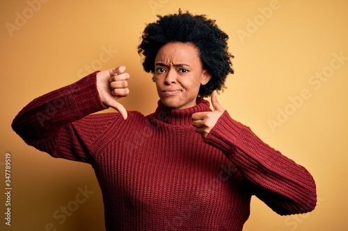 Young beautiful African American afro woman with curly hair wearing casual turtleneck sweater Doing thumbs up and down, disagreement and agreement expression. Crazy conflict