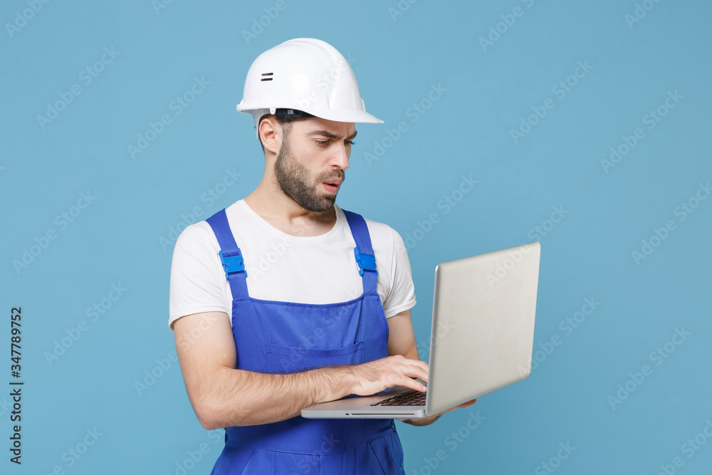 Worried young man in coveralls protective helmet hardhat working on laptop pc computer isolated on blue background studio. Instruments accessories for renovation apartment room. Repair home concept.