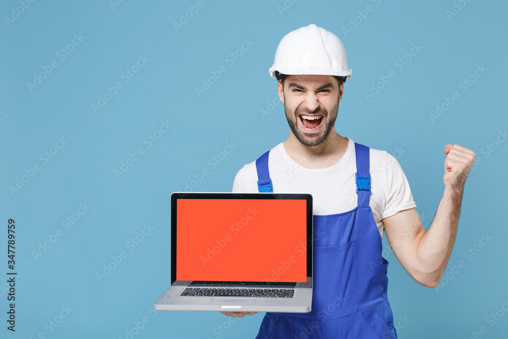 Happy man in coveralls hardhat hold laptop computer with blank empty screen isolated on blue background. Instruments accessories renovation apartment room. Repair home concept. Doing winner gesture. Happy man in coveralls hardhat hold laptop computer with blank empty screen isolated on blue background. Instruments accessories renovation apartment room. Repair home concept. Doing winner gesture.
