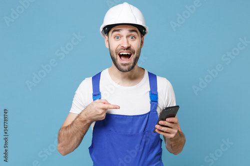 Shocked young man in coveralls protective helmet hardhat pointing index finger on mobile phone isolated on blue background. Instruments accessories for renovation apartment room. Repair home concept.