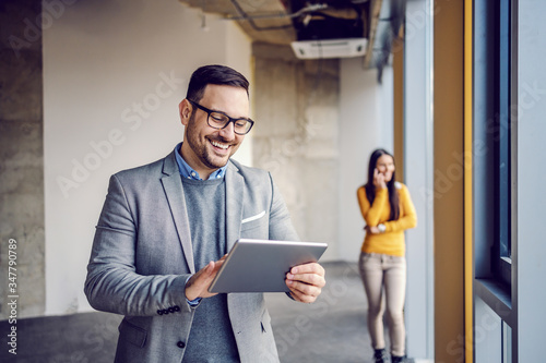 Handsome real estate agent in suit standing in building in construction process and using tablet for make appointment with customers. In background his colleague standing and talking on the phone.