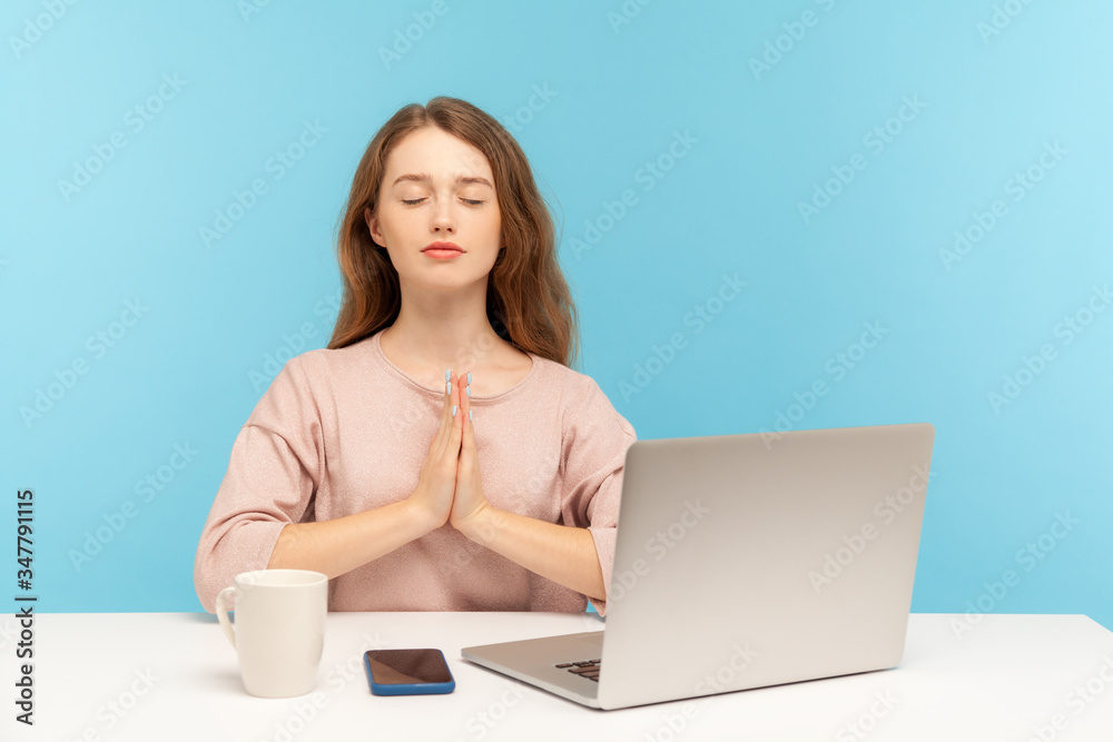 Break at work, yoga practice. Calm woman employee sitting at workplace with laptop and raising hands in namaste gesture, meditating at home office. indoor studio shot isolated on blue background Break at work, yoga practice. Calm woman employee sitting at workplace with laptop and raising hands in namaste gesture, meditating at home office. indoor studio shot isolated on blue background