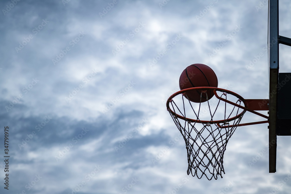 Dark silhouette of street basketball ball falling into the hoop on a cloudy day. Close up of a ball above the hoop net. Concept of success, scoring points and winning. Urban youth game.