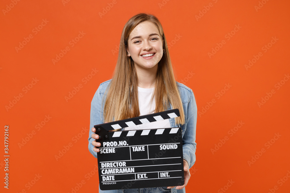 Smiling young woman girl in casual denim clothes posing isolated on orange wall background studio portrait. People lifestyle concept. Mock up copy space. Hold classic black film making clapperboard.
