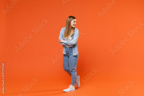 Side view of cheerful young woman girl in casual denim clothes posing isolated on orange background studio portrait. People lifestyle concept. Mock up copy space. Holding hands crossed looking aside.