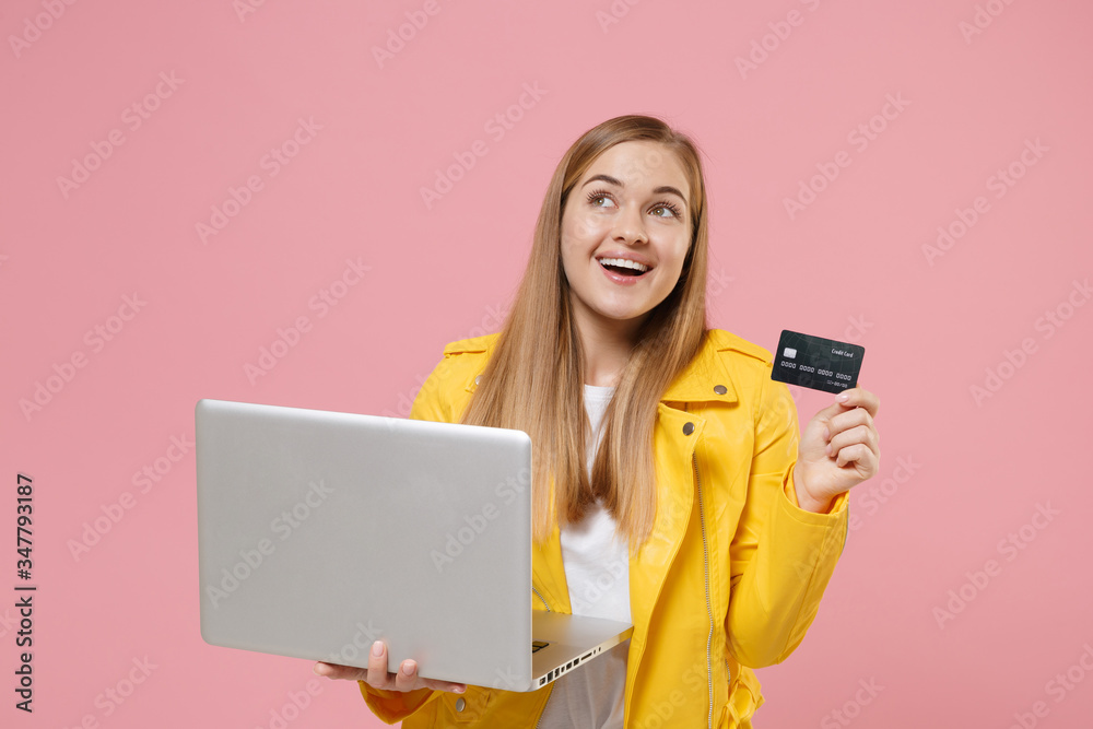 Funny young woman girl in yellow leather jacket posing isolated on pastel pink background studio. People lifestyle concept. Mock up copy space. Hold laptop pc computer credit bank card, looking up. Funny young woman girl in yellow leather jacket posing isolated on pastel pink background studio. People lifestyle concept. Mock up copy space. Hold laptop pc computer credit bank card, looking up.