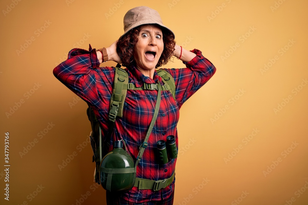Middle age curly hair hiker woman hiking wearing backpack and water canteen using binoculars Crazy and scared with hands on head, afraid and surprised of shock with open mouth