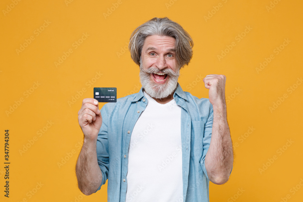 Joyful elderly gray-haired mustache bearded man in casual blue shirt posing isolated on yellow background. People lifestyle concept. Mock up copy space. Hold credit bank card, doing winner gesture. Joyful elderly gray-haired mustache bearded man in casual blue shirt posing isolated on yellow background. People lifestyle concept. Mock up copy space. Hold credit bank card, doing winner gesture.