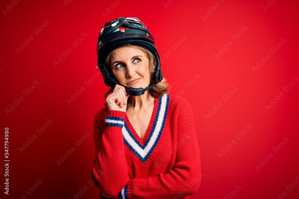 Middle age beautiful blonde motorcyclist woman wearing moto helmet over red background with hand on chin thinking about question, pensive expression. Smiling with thoughtful face. Doubt concept. Middle age beautiful blonde motorcyclist woman wearing moto helmet over red background with hand on chin thinking about question, pensive expression. Smiling with thoughtful face. Doubt concept.