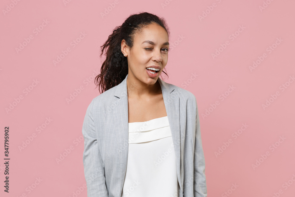 Fototapeta premium Cheerful funny young african american business woman in grey suit white shirt posing isolated on pink background in studio. Achievement career wealth business concept. Mock up copy space. Blinking.