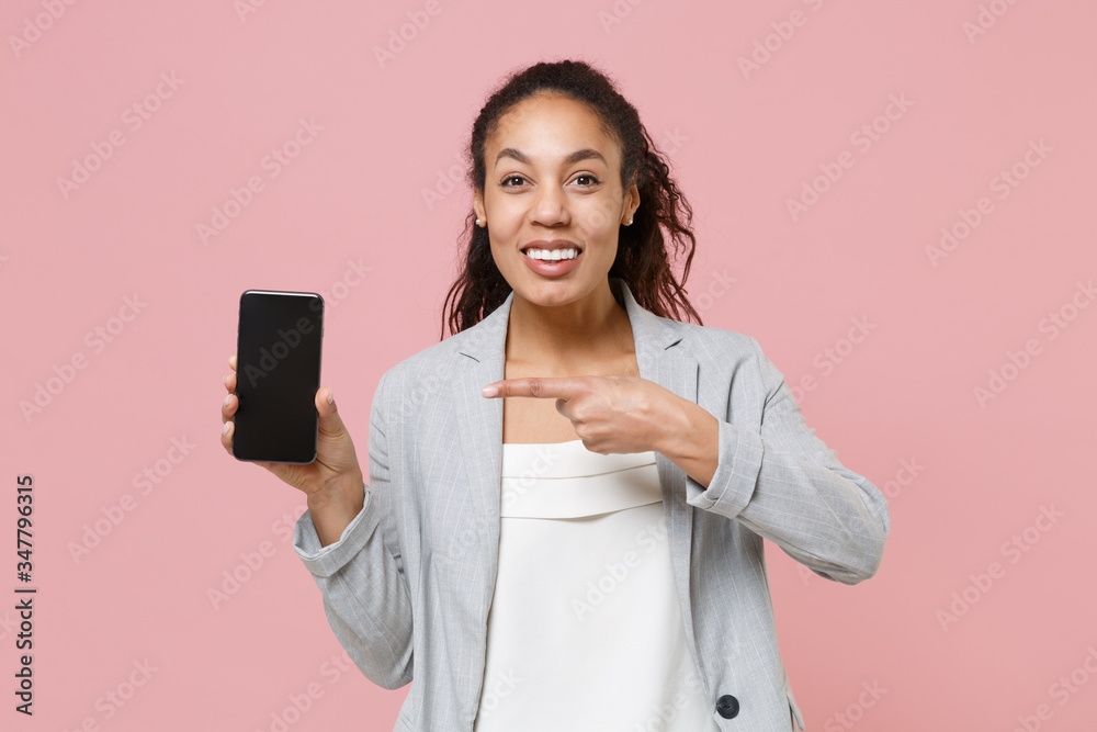 Smiling african american business woman in grey suit white shirt isolated on pink background. Achievement career wealth business concept. Pointing index finger on mobile phone with blank empty screen. Smiling african american business woman in grey suit white shirt isolated on pink background. Achievement career wealth business concept. Pointing index finger on mobile phone with blank empty screen.