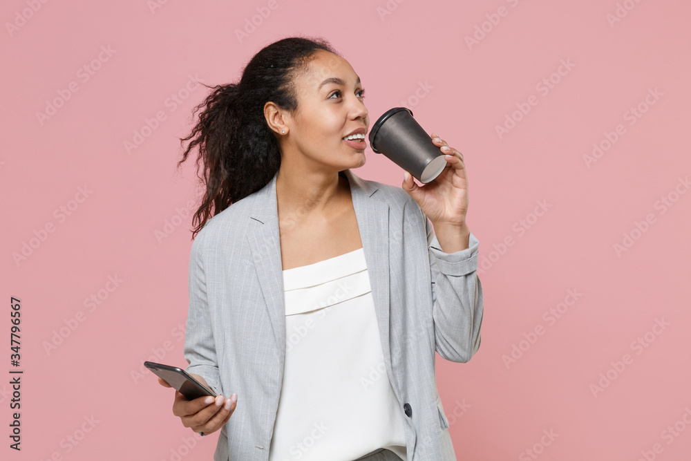 Beautiful young african american business woman in grey suit white shirt isolated on pink background. Achievement career wealth business concept. Using mobile phone, hold paper cup with coffee or tea. Beautiful young african american business woman in grey suit white shirt isolated on pink background. Achievement career wealth business concept. Using mobile phone, hold paper cup with coffee or tea.