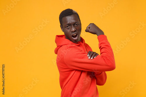 Side view of young african american man guy in red streetwear hoodie posing isolated on yellow wall background studio portrait. People lifestyle concept. Mock up copy space. Showing biceps muscles.
