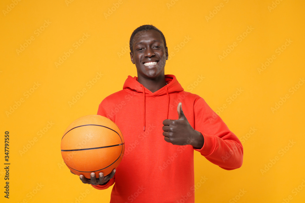 Smiling young african american man guy basketball player in red streetwear hoodie posing isolated on yellow background. Sport leisure lifestyle concept. Playing basketball hold ball showing thumb up. Smiling young african american man guy basketball player in red streetwear hoodie posing isolated on yellow background. Sport leisure lifestyle concept. Playing basketball hold ball showing thumb up.